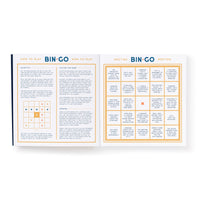 An open Bin-go To A Dumb Meeting Bingo Book by Brass Monkey shows play instructions on the left and a vibrant Bingo card with yellow and blue meeting scenarios on the right.