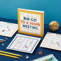 A desk with Brass Monkeys Bin-go To A Dumb Meeting Bingo Book, yellow and white box, plus paper clips, a rubber band ball, pens, pencils, and notepads—fun office essentials set against a blue background.