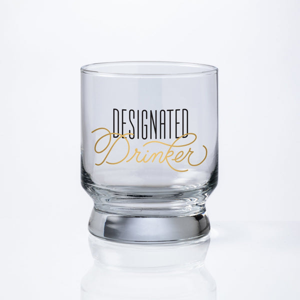 The Brass Monkey Designated Drinker Lowball Glass features Designated Drinker in black and gold lettering, shown stackable on a plain white background.