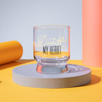 The Distill My Heart Lowball Glass by Brass Monkey rests on a round, light purple platform with orange and yellow cylinders and a yellow surface in the background.