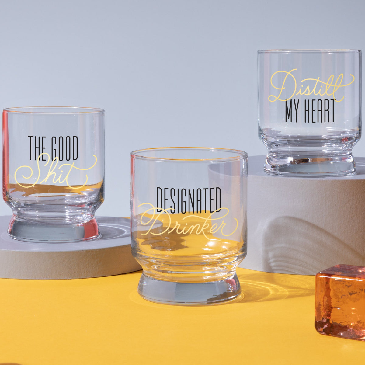 Three stackable Brass Monkey Distill My Heart Lowball Glasses sit on gray platforms against a yellow backdrop, featuring playful text—The Good Shit, Designated Drinker—and bold black and yellow artwork.