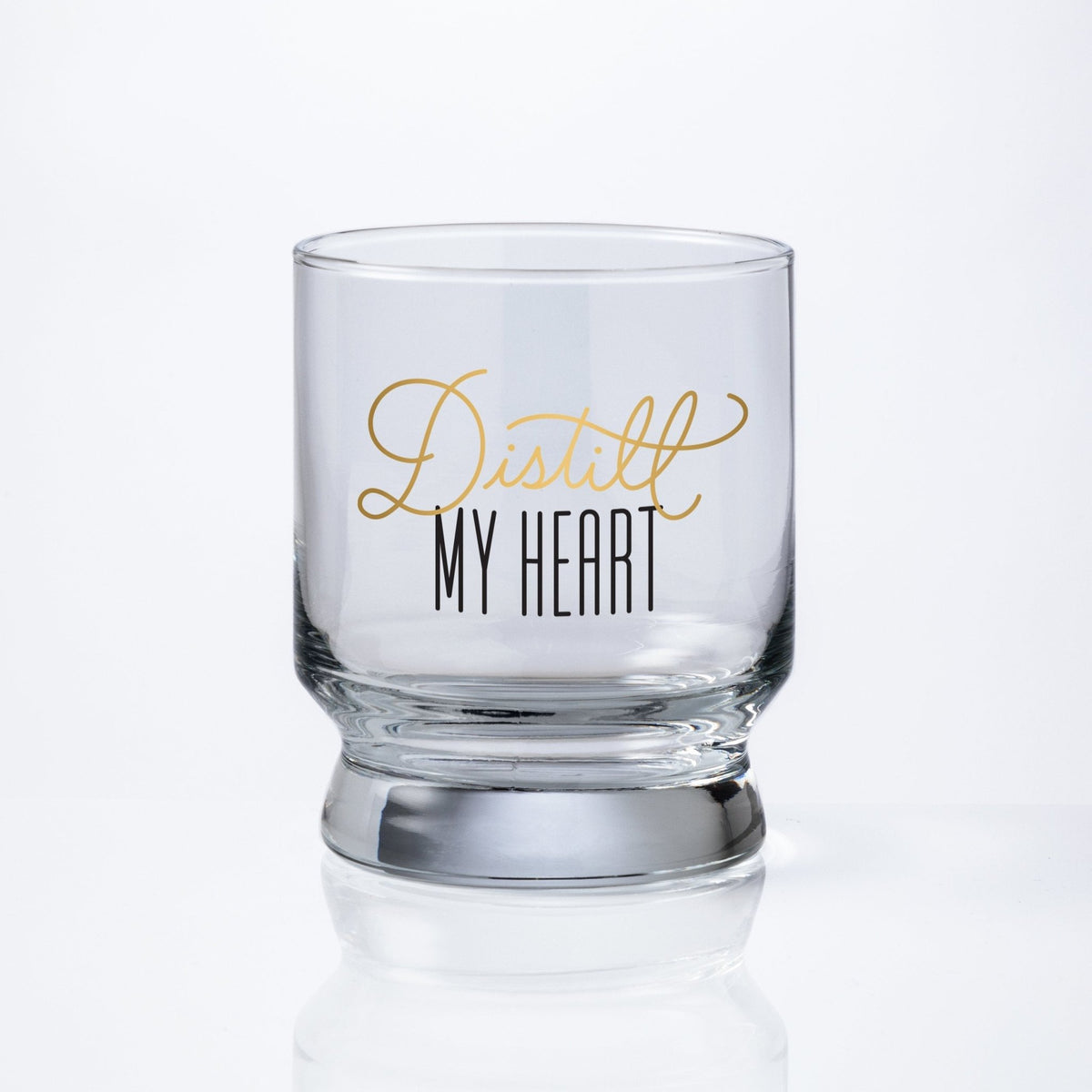 The Brass Monkey Distill My Heart Lowball Glass is a clear, pedestal-shaped glass with Distill My Heart in gold and black script on the front, set against a white background for an elegant, sophisticated look.