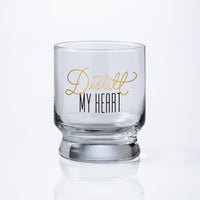 The Brass Monkey Distill My Heart Lowball Glass is a clear, pedestal-shaped glass with Distill My Heart in gold and black script on the front, set against a white background for an elegant, sophisticated look.