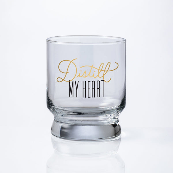 The Brass Monkey Distill My Heart Lowball Glass is a clear, pedestal-shaped glass with Distill My Heart in gold and black script on the front, set against a white background for an elegant, sophisticated look.