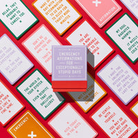 A Brass Monkey Emergency Affirmations for Exceptionally Stupid Days Card Deck is displayed on a bright red surface, surrounded by the brands colorful affirmation cards featuring humorous and encouraging messages.