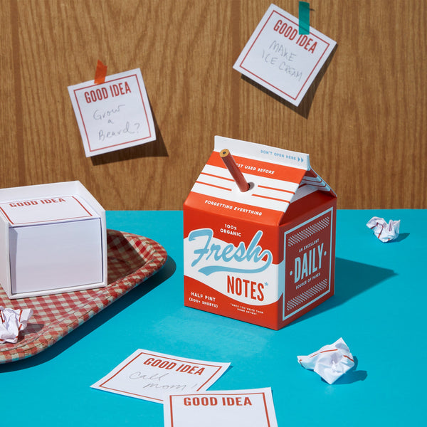 A vibrant scene featuring a red and blue Fresh Ideas Milk Carton Note Set by Brass Monkey, surrounded by Good Idea cards, crumpled papers, and a checkered tray on a wooden backdrop.