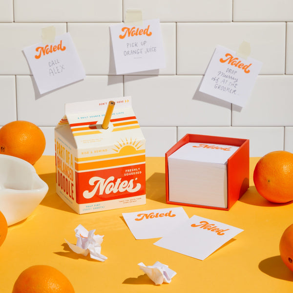Brass Monkey’s Freshly Squeezed Notes notepad block, shaped like an orange juice carton, sits among oranges and notes with a pencil in the straw hole, while sticky notes are displayed on a tiled wall.