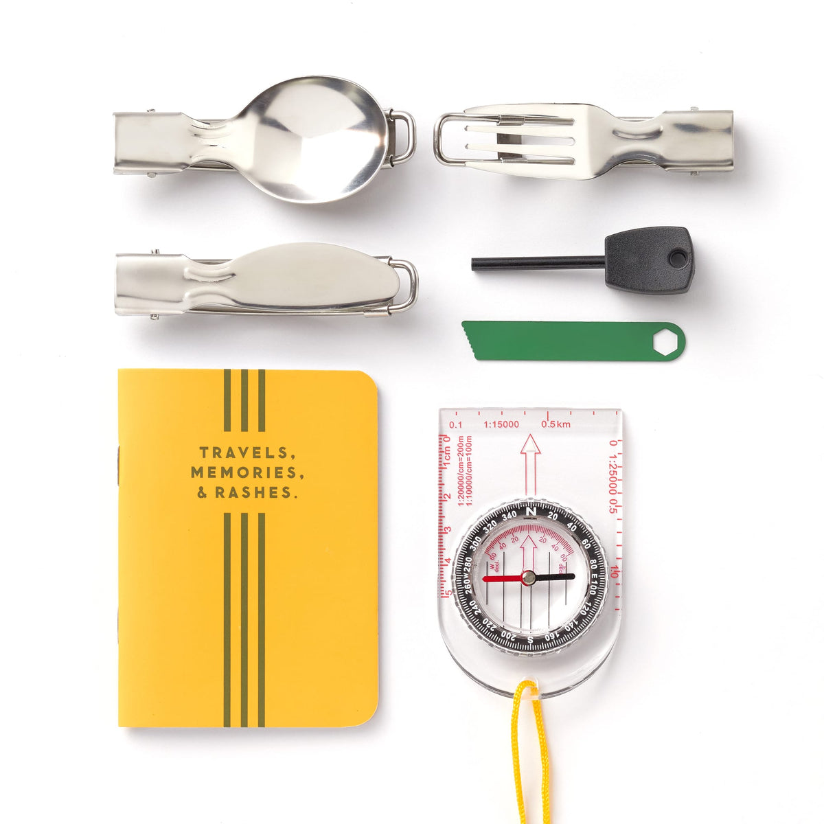 A flat lay of the Brass Monkey Get Lost Camping Survival Kit, featuring a foldable spoon, fork, knife, fire starter, green multitool, survival items, yellow TRAVELS journal, and a compass with measuring tool and yellow string.