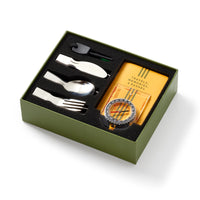 The Brass Monkey Get Lost Camping Survival Kit features a green box containing a silver spork, knife, spoon, black whistle, yellow journal marked Travels, Memories, Dreams, and a black compass with yellow accents—all in black foam.