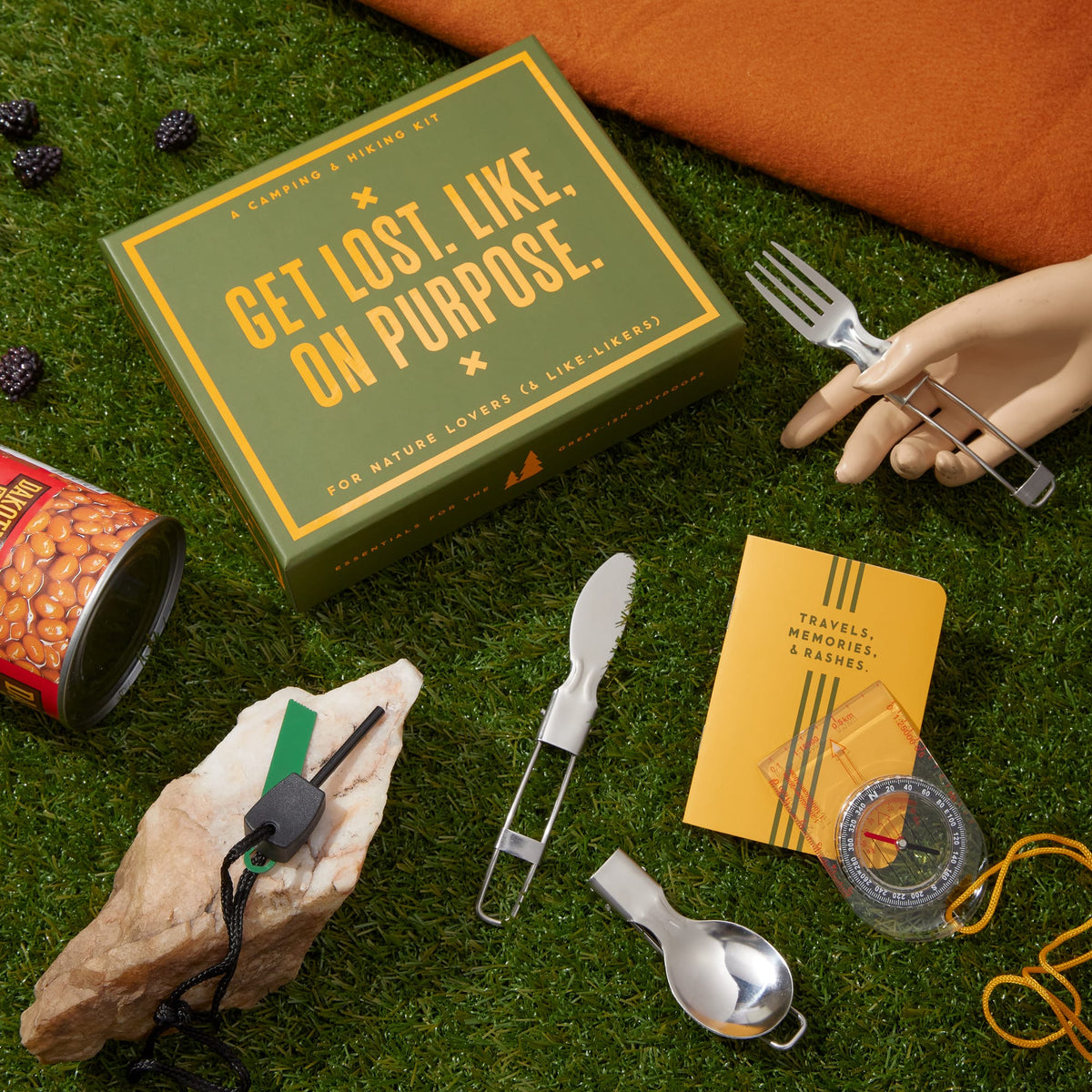The Brass Monkey Get Lost Camping Survival Kit rests on grass, featuring a yellow box, can of beans, penknife, metal utensils, compass, travel journal notebook, mannequin hand with fork; blackberries and an orange cloth are nearby.