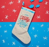 The Here Kitty Kitty Cat Stocking by Brass Monkey features cream velvet, red and blue Here Kitty Kitty embroidery, blue felt fish pieces, and is displayed on a festive red and blue Brass Monkey background.