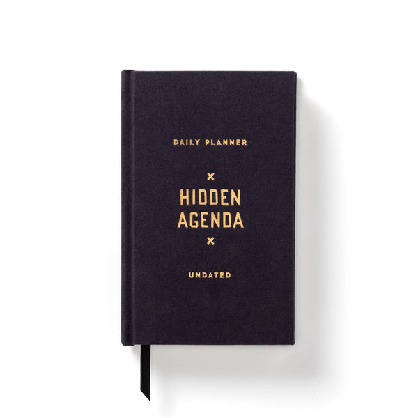 The Brass Monkey Hidden Agenda Undated Mini Planner is a black hardcover vintage-inspired planner with gold text on the cover and a black ribbon bookmark.