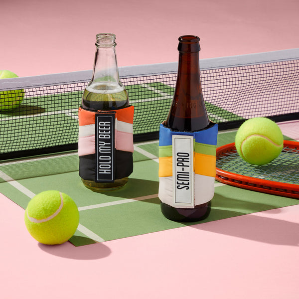 Two beer bottles in Brass Monkey’s Hold My Beer Puffyvest Coozie Set, labeled HOLD MY BEER and SEMI-PRO, rest on a toy tennis court with a red racket and three tennis balls against a playful pink background.