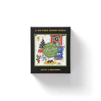 The Brass Monkey Home For Christmas 100 Piece Mini Shaped Puzzle comes in a giftable box featuring a festive living room scene with a tree, gifts, and cat. The cover says Ill be home for Christmas and has space for your personal message.