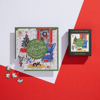 The completed Brass Monkey Home For Christmas 100 Piece Mini Shaped Puzzle, featuring pets by a fireplace and tree, sits on a white and red background with four loose pieces and its giftable box displaying the same festive image nearby.