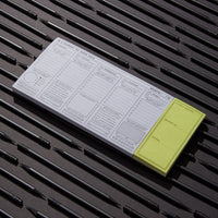 The Brass Monkey In a Moment of Weekness Weekly Planner Pad, vintage-inspired with daily planning sections and a yellow sticky note, is arranged on a surface with evenly spaced black pens, its organized layout emphasized by sharp lighting.