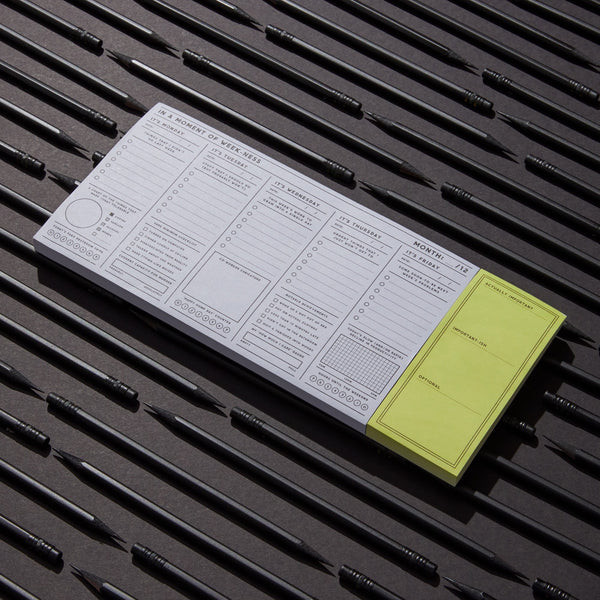 The Brass Monkey In a Moment of Weekness Weekly Planner Pad, vintage-inspired with daily planning sections and a yellow sticky note, is arranged on a surface with evenly spaced black pens, its organized layout emphasized by sharp lighting.