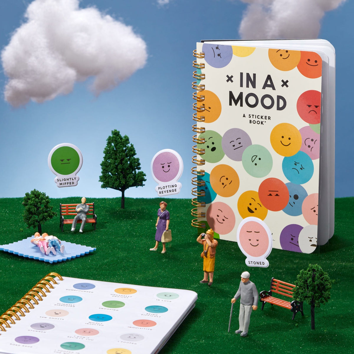 The In A Mood Sticker Book by Brass Monkey stands upright on artificial grass with miniature figurines, trees, and benches. Its cover displays colorful moody emoticons, with expression stickers visible nearby.