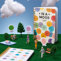 The In A Mood Sticker Book by Brass Monkey stands upright on artificial grass with miniature figurines, trees, and benches. Its cover displays colorful moody emoticons, with expression stickers visible nearby.