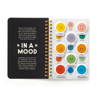 The Brass Monkey In A Mood Sticker Book features an open spiral notebook with a black page titled IN A MOOD and twelve stickers on the right, each showing moody emoticons labeled with emotions like FEELING FLIRTY and KINDA MURDERY.