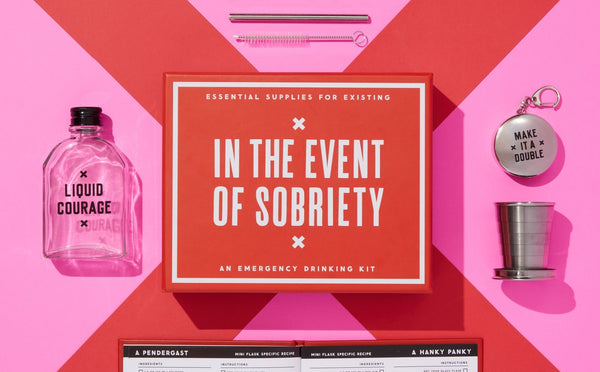 A pink background features a red Brass Monkey In The Event Of Sobriety Emergency Drinking Kit box in the center, surrounded by a flask, collapsible travel cup, jigger, keychain, and mini cocktail recipe book.