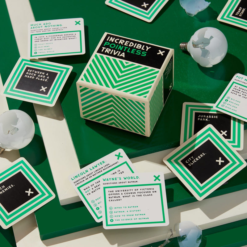 A box of Brass Monkey’s Incredibly Pointless Trivia rests on a green surface, surrounded by question cards with categories like Wayne’s World, Lincoln Lawyers, and Jurassic Park, plus several broken light bulbs.