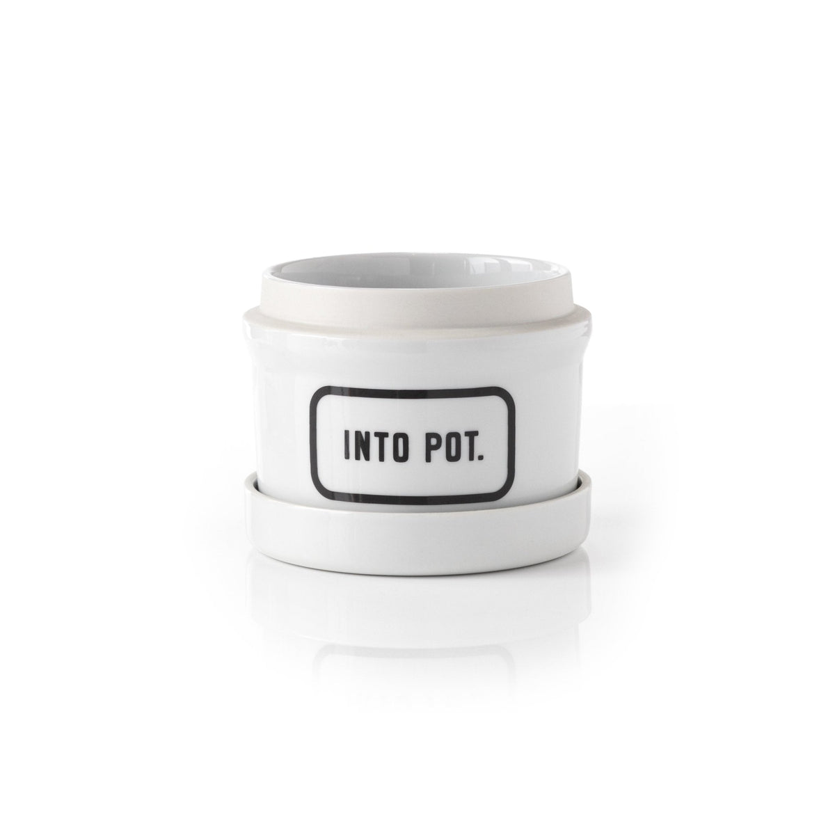 The Brass Monkey Into Pot Planter w/ Drip Tray is a white ceramic planter with a matching saucer and a bold, black-bordered “INTO POT.” label on the front, combining vintage style and modern functionality.