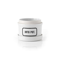 The Brass Monkey Into Pot Planter w/ Drip Tray is a white ceramic planter with a matching saucer and a bold, black-bordered “INTO POT.” label on the front, combining vintage style and modern functionality.