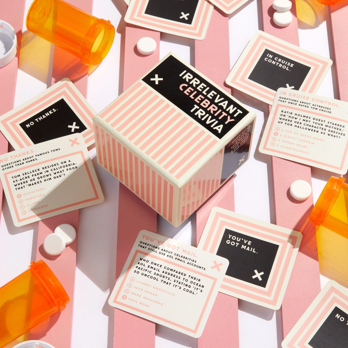 A box of Brass Monkeys Irrelevant Celebrity Trivia rests on a pink-and-white striped surface, surrounded by game cards and orange bottles. The cards offer fun celebrity trivia—ideal for anyone who loves quirky pop culture facts.