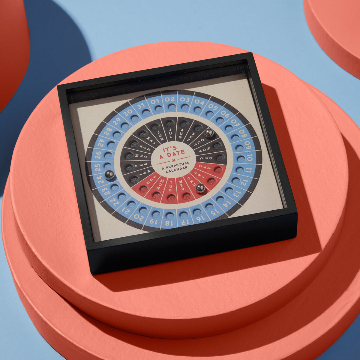 The Brass Monkey Its A Date Perpetual Desk Calendar features rotating rings to display dates, days, and months. Stainless steel balls accent the square design on a round peach platform, set against a light blue and peach background.
