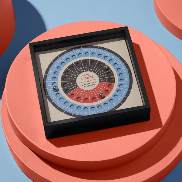 The Brass Monkey Its A Date Perpetual Desk Calendar features rotating rings to display dates, days, and months. Stainless steel balls accent the square design on a round peach platform, set against a light blue and peach background.