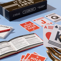 The Brass Monkey Its In The Cards Playing Card Game Set includes oversized cards, a scorebook, red Hold My Hand cards, a rulebook, and comes in a sleek black box—ideal for fans of card games.