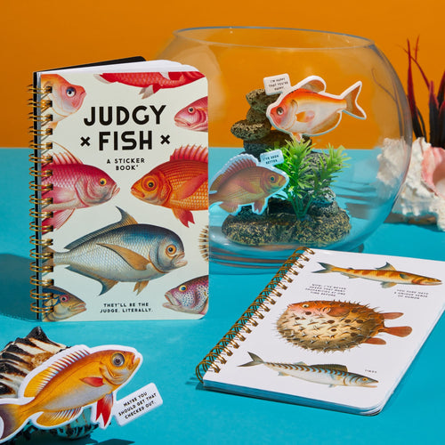 The Brass Monkey Judgy Fish Sticker Book, with vintage fish on the cover, sits alongside a spiral notebook and fishbowl with quirky stickers, all arranged on a blue surface against an orange backdrop.