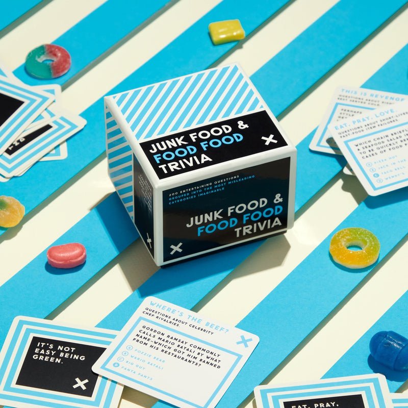 A cube-shaped Brass Monkey Junk Food & Food Food Trivia box sits on a striped surface, surrounded by colorful gummy candies and trivia cards featuring fun food quiz questions and answers.