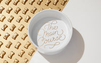 The Main Course Pet Food Bowl by Brass Monkey, a sturdy ceramic dish with reflective gold lettering, is displayed beside a gold mat lined with shiny bone-shaped dog treats.