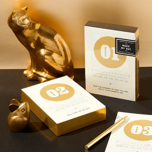 A stylish setup featuring Brass Monkeys Make My Day Undated Desk Calendar, a vintage-inspired gold cat and mouse figurine, gold pen, and two boxed stationery sets labeled 01 and 02 on a beige and gold backdrop.