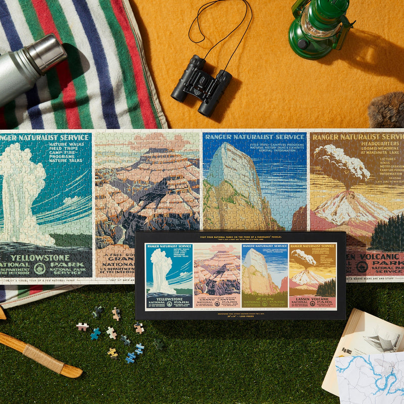 The Brass Monkey National Parks 1,000 Piece Panoramic Puzzle, inspired by vintage park posters, is completed on grass beside its box, a striped blanket, binoculars, lantern, thermos, spare puzzle pieces, and a map—perfect for adventure lovers.