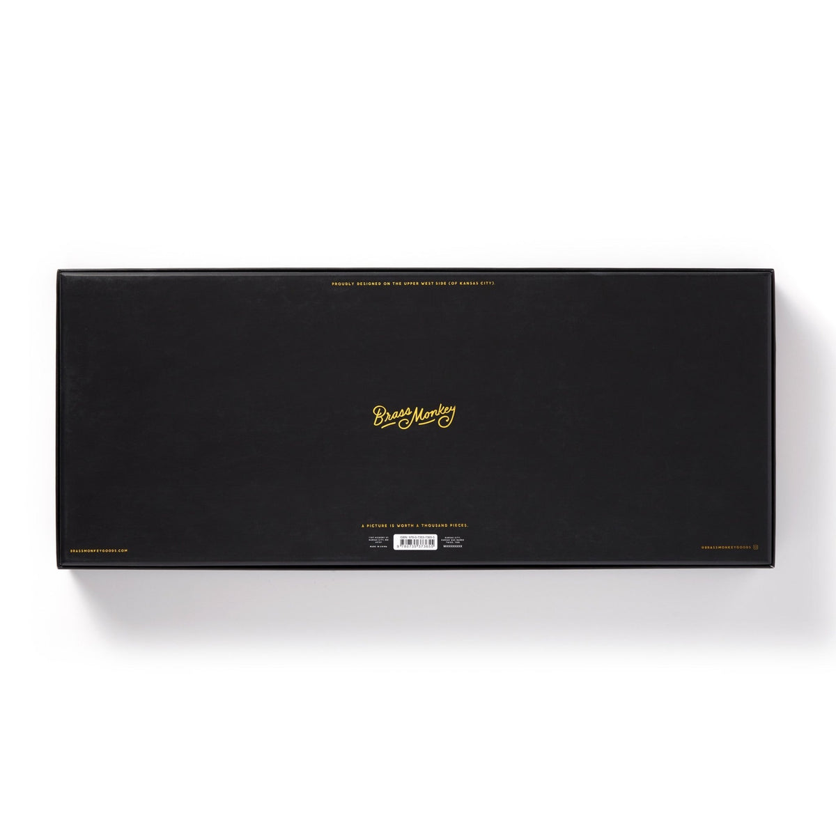 A large rectangular black box with gold text that reads “NYC Map 1,000 Piece Panoramic Puzzle” by Brass Monkey in the center and small gold writing along the edges, resembling a vintage map, photographed on a white background.