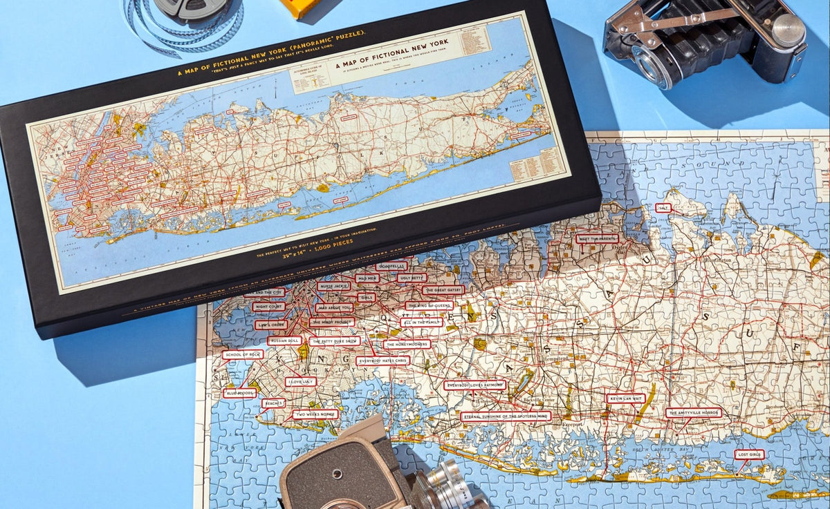 A partially completed Brass Monkey NYC Map 1,000 Piece Panoramic Puzzle rests on a blue surface, featuring red-labeled place names. Nearby are a vintage map of New York City, film reel, retro camera, and lens.
