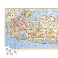 The NYC Map 1,000 Piece Panoramic Puzzle by Brass Monkey shows a vintage map of New York City with place names in red rectangles and loose pieces in the lower left corner.