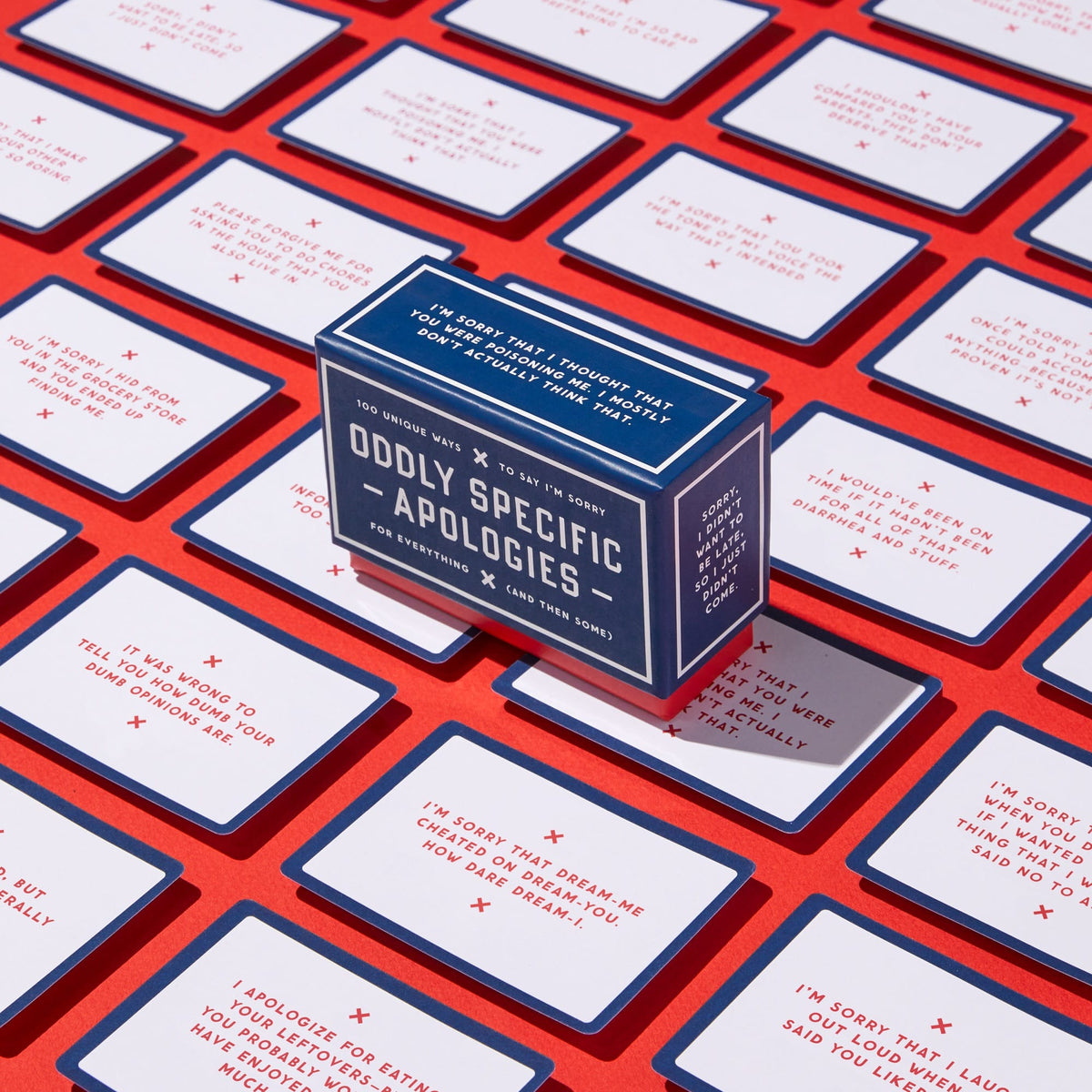 A blue portable box labeled Oddly Specific Apologies by Brass Monkey sits on a red surface, surrounded by unique apology cards featuring humorous, specific messages in red and blue text.