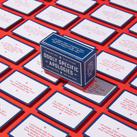 A blue portable box labeled Oddly Specific Apologies by Brass Monkey sits on a red surface, surrounded by unique apology cards featuring humorous, specific messages in red and blue text.