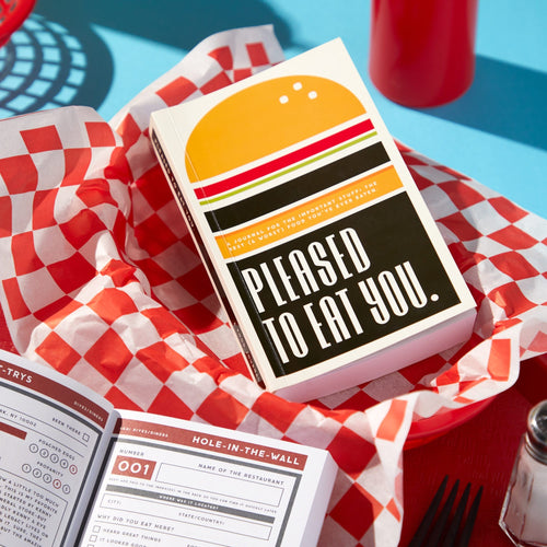 The Pleased to Eat You Food Journal by Brass Monkey, with a burger-themed cover, sits in a red plastic basket lined with checkered paper beside an open restaurant logbook and a bright red ketchup bottle.
