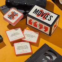 Brass Monkeys Poorly Explained Movies Game is shown with its box, cards featuring quirky movie plot summaries, and two VHS tapes, all arranged on a bright orange surface.