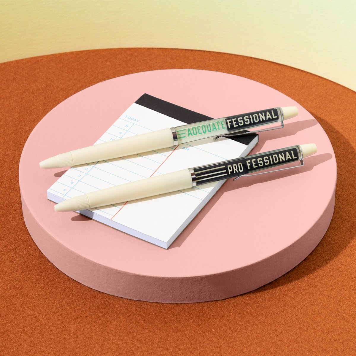 Two cream-colored pens from Brass Monkey’s Professional Procrastinator Floaty Pen Set, labeled ADEQUATE FESSIONAL and PRO FESSIONAL, rest on a lined checklist notepad atop a pink round platform with a brown and yellow background.