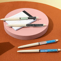 The Brass Monkey Professional Procrastinator Floaty Pen Set includes four white ballpoint pens with humorous labels, arranged on a pink platform and brown surface, with a small notepad in the background.