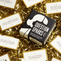 A box of Brass Monkey’s Question Sparks rests on gold tinsel, surrounded by cards with fun conversation starters and questions printed in black on white backgrounds.