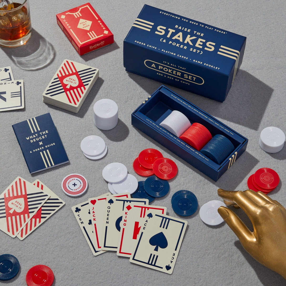 The Raise The Stakes Poker Game Set by Brass Monkey features a deck of cards, red, white, and blue poker chips, all in a branded box—displayed with a drink and gold mannequin hand on a gray surface.