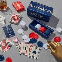 The Raise The Stakes Poker Game Set by Brass Monkey features a deck of cards, red, white, and blue poker chips, all in a branded box—displayed with a drink and gold mannequin hand on a gray surface.