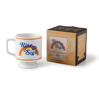 The Brass Monkey Rise & Sigh Pedestal Mug, a white stackable ceramic mug with a rainbow and vintage charm, sits beside its matching brown box packaging, both featuring the same unique “Rise & Sigh” design.